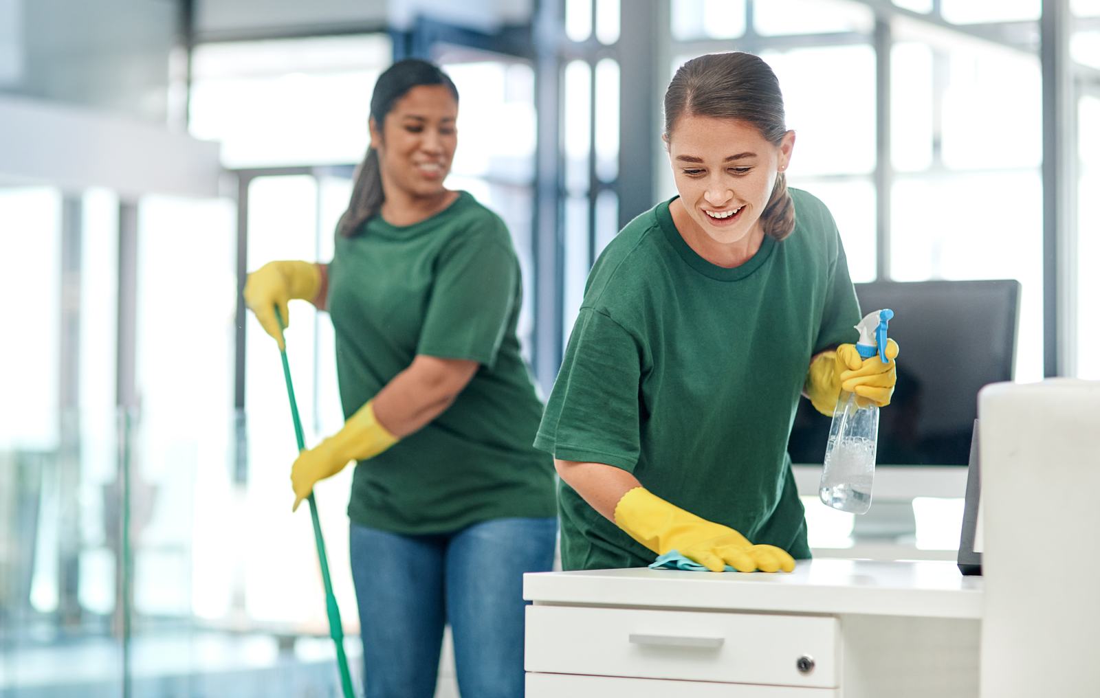 Two custodians in green shirts cleaning an office desk and floor